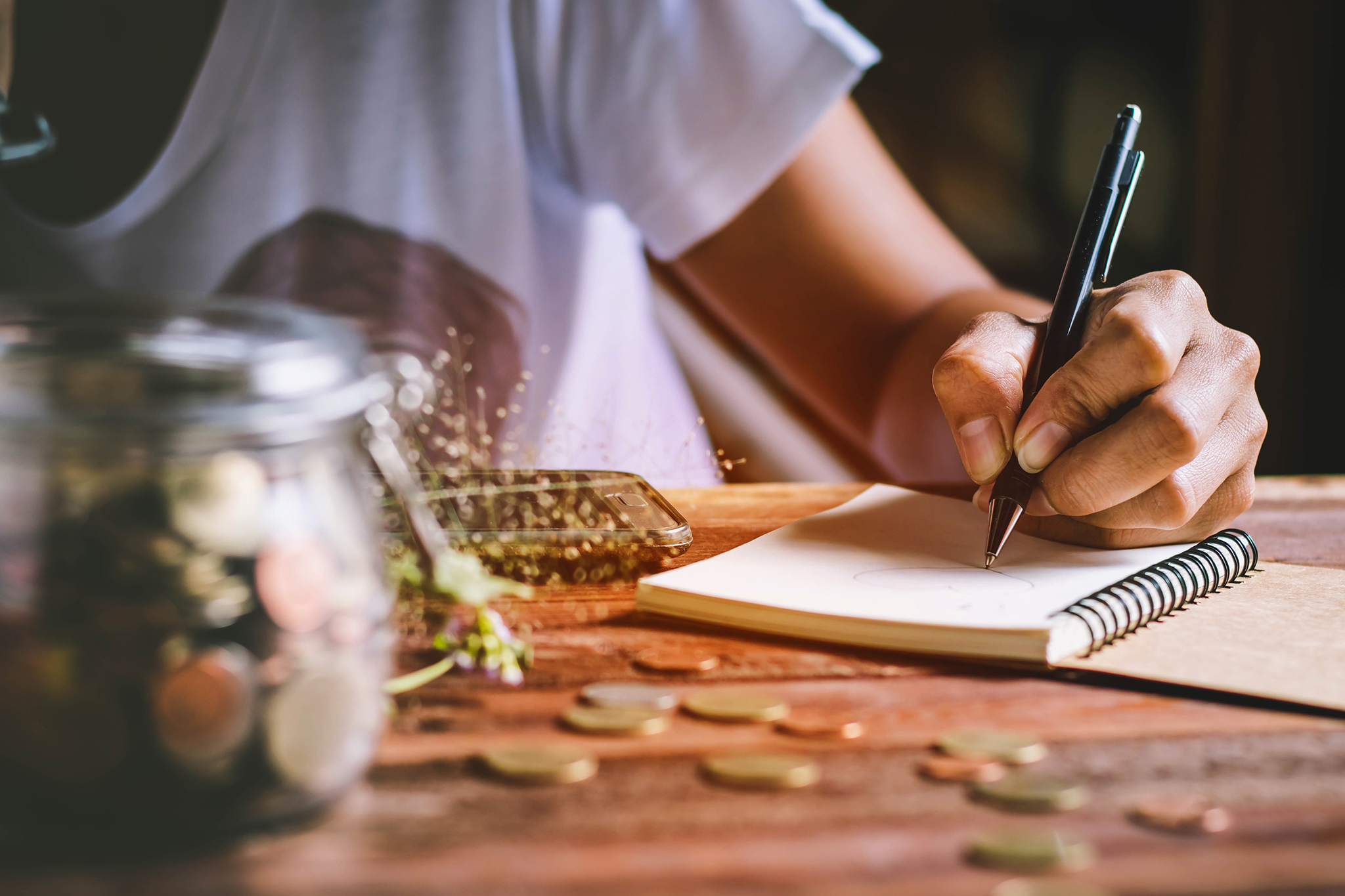 Someone sat at a table writing notes on a notepad with a jar of coins beside them