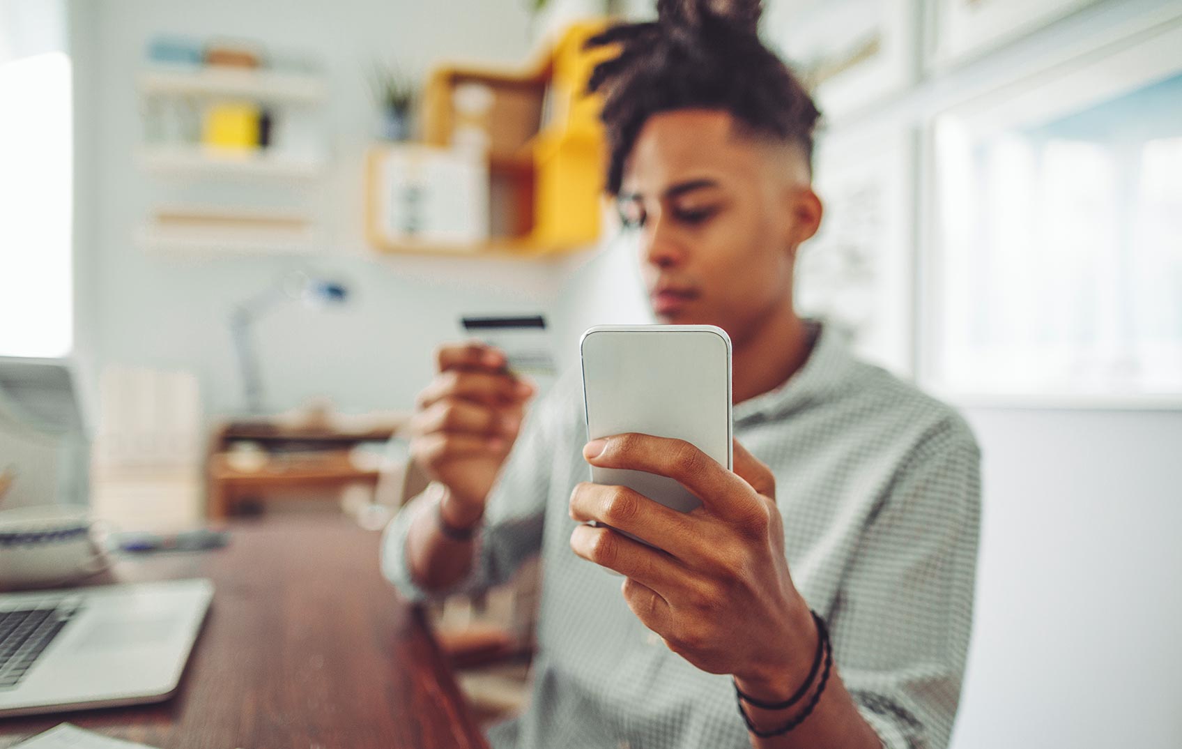 A male student sits at his laptop whilst holding up his mobile and his bank card to make an online payment